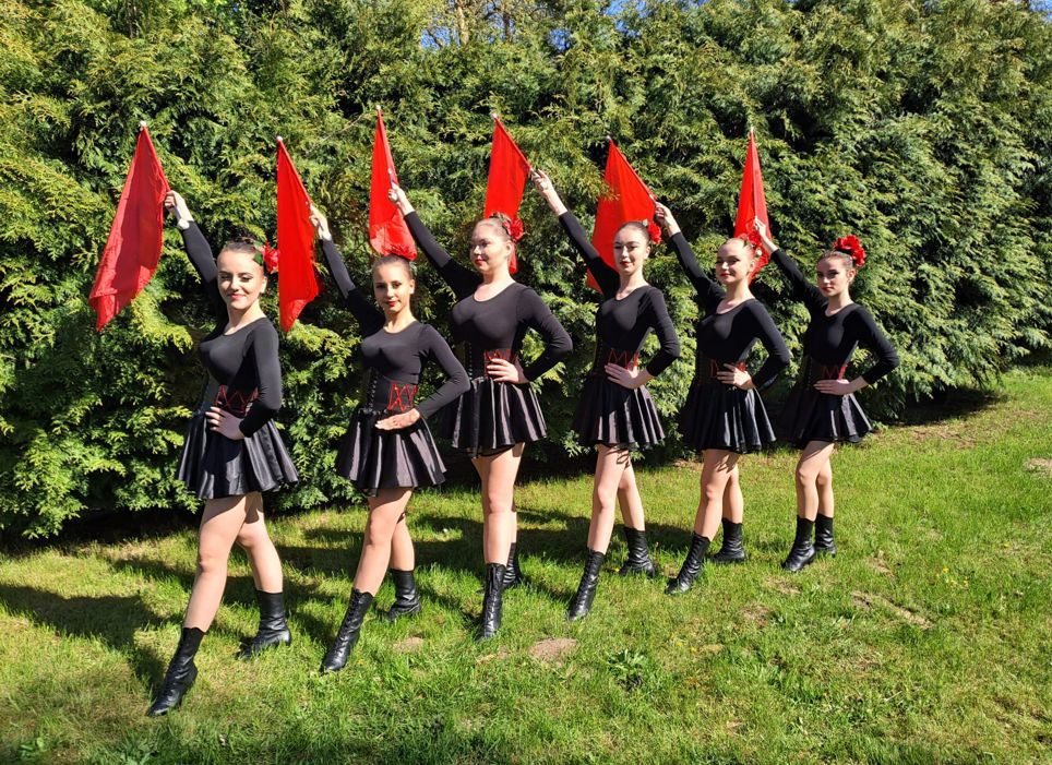 sześć dziewcząt w szeregu z flagami podniesionymi do góry i lewą logą w wykroku, w krótkich czarnych sukienkach na tle tui